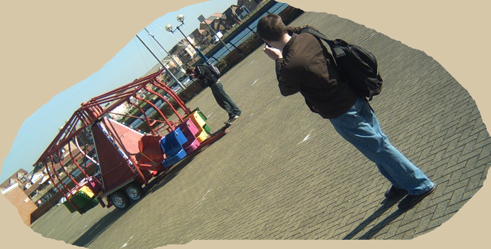 Shot at an angle of about 45 degrees, a paved area of Hartlepool marina, with a quay and flats in the background. In the foreground, Martyn photographs Gary, a short distance away, who photographs a detail on a small, colourful, inactive fairground ride. The scene has been cut around digitally, in an irregular freehand shape, and the area outside is filled with a flat light tan colour.