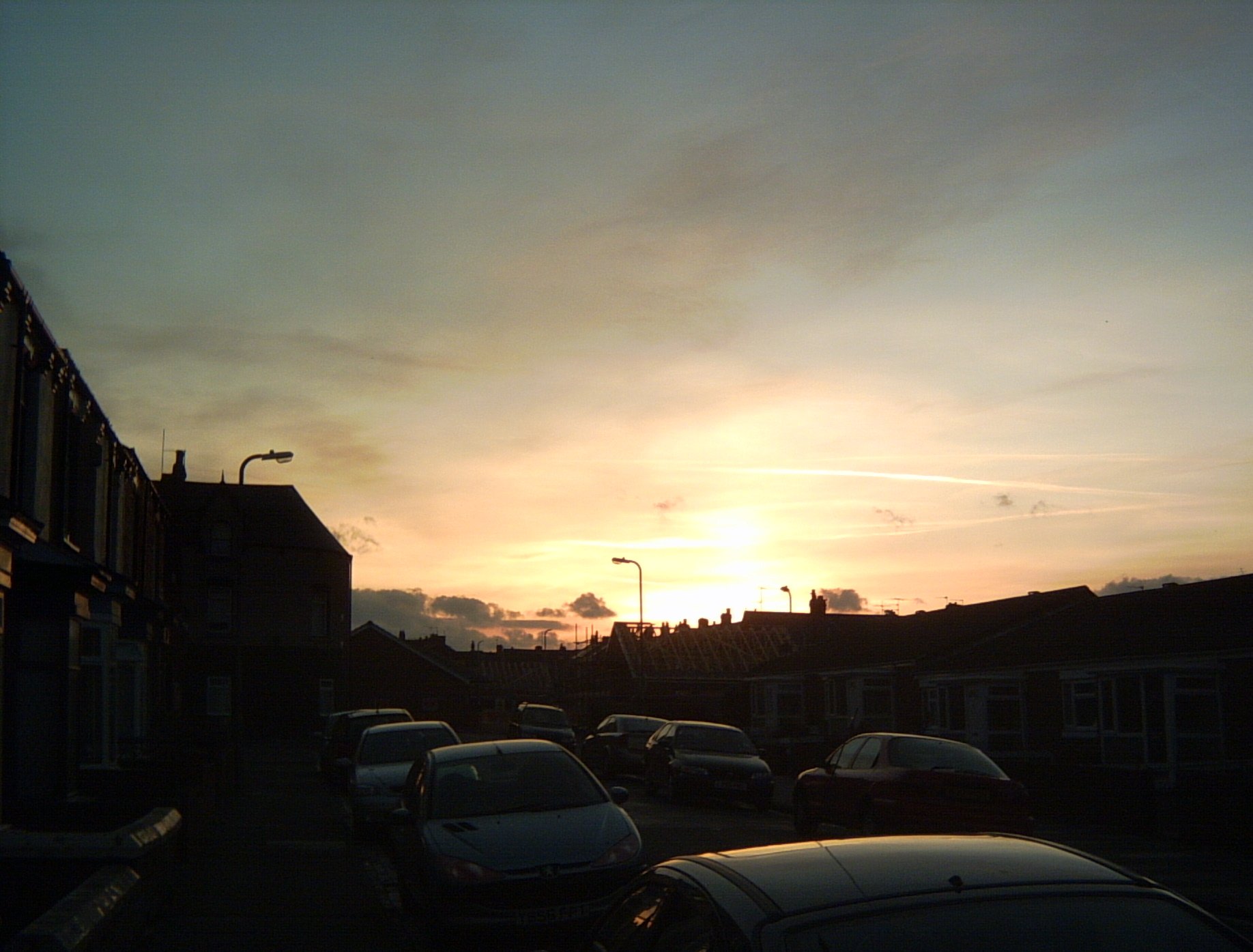 A sunset over a residential street of two-storey houses, viewed along the street, which is lined with parked cars. There are a few heavy clouds in the distance, and lighter clouds texturing the sky. The sky is yellow at the centre, behind a protruding streetlight immediately above the rooftops, and fades through pale peach to duck-egg blue at the edges.