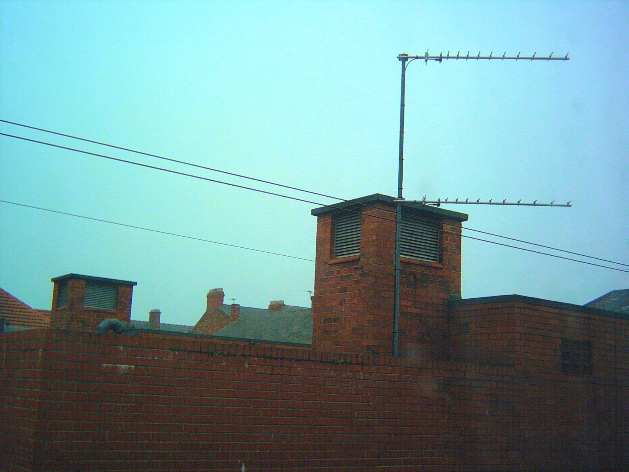 The top of a reddish-brown brick building, a square wall, with no visible rooftop. A corner is just in view to the left; the left side wall extends away out of the image, and the front wall falls gradually away throughout, to the right of the image. Just right of centre, protruding above the building's front wall is a small square tower viewed diagonally, about a metre tall like a chimney without chimneypots, and with a metal slatted grate at the top of each of the 2 visible side faces. On top of the front wall in front of the tower is a metal pole extending about 2 metres, with an aerial half-way up and another at the top, both pointing right, like an “F”. A pair of wires passes through the whole image in front of the pole and tower, parallel to the top of the front wall; another parallel wire ends attached to the pole. The sky is blue with a yellowish tint.