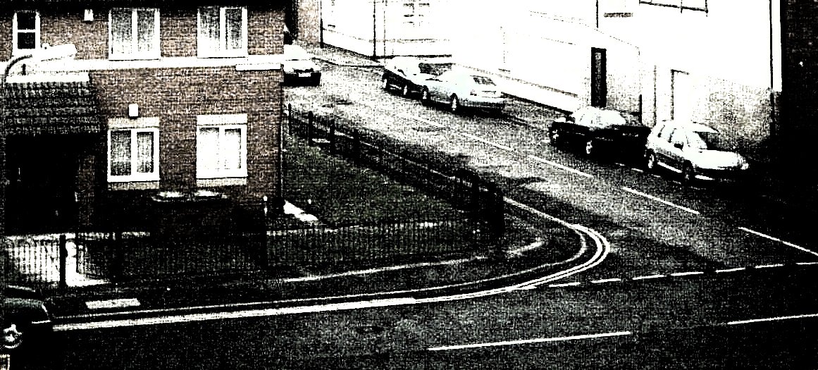 A junction of 2 residential streets: a house with a small front garden and low metal fence, and another street extending away behind it. There are a few parked cars. The image is grainy and dithered, like a low-quality ink printing on cheap zine paper. The colours are washed out and almost completely desaturated: the house's bricks look very faintly brown, and its grass looks like it might be dark green; the houses on the other street all appear white with indistinct outlines of doors and windows; the road surface is black with sharp white lines; the cars appear black white and grey, but could be any colour.
